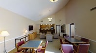 This single-story clubhouse offers a welcoming, multi-functional common area designed for community gatherings and everyday social interaction. The space is immediately distinguished by its soaring vaulted ceilings, which lend an open, airy quality to the interior and are complemented by warm beige walls throughout.

The main area is thoughtfully arranged into distinct zones. A central lounge area features upholstered armchairs and soft seating arranged around a low table, creating an inviting conversation space. Nearby, several dining tables paired with wooden and cushioned chairs provide ample seating for meals or group activities. An elegant chandelier and recessed lighting ensure the space is well-lit throughout.

Toward the back, a fully equipped kitchenette with wooden cabinetry, counter seating, and appliances supports food preparation and serving for events. A flat-screen television mounted on the wall adds an entertainment element to the lounge area, while a side console with a lamp contributes warm, ambient lighting.

Natural light filters into the space, enhancing the overall brightness. The layout balances comfort and functionality, making this clubhouse a versatile hub for community life.