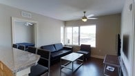 This single-floor space presents a clean, modern aesthetic centered around a well-appointed living area. The room features dark hardwood flooring that contrasts with neutral-toned walls, creating a contemporary and cohesive look throughout. A ceiling fan with a built-in light fixture is centrally mounted, providing both air circulation and ambient lighting to the space.

The living area is furnished with a black leather sofa and a matching accent chair, complemented by a sleek metal and glass coffee table. Two large windows allow generous natural light to fill the room while offering views of the surrounding outdoor environment, enhancing the sense of openness despite the compact scale.

Adjacent to the living area, a dining space is partially visible, featuring dark chairs and a granite-topped surface, suggesting a practical and stylish transition between the living and dining zones. A mirrored surface near the entryway adds depth and reflects additional light into the space.

Overall, the unit maintains a straightforward, functional layout with modern finishes, making it a practical and visually appealing living environment suited for everyday comfort.