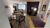 This single-floor space showcases a thoughtfully designed interior that balances comfort and contemporary style. The living area is anchored by classic parquet hardwood flooring, which adds warmth and texture throughout. A striking deep purple accent wall serves as a bold focal point, contrasting elegantly against the crisp white walls that define the rest of the space.

The seating area is furnished with a neutral-toned sofa adorned with decorative cushions in complementary earth tones and purples, paired with a patterned accent chair featuring a geometric design. A glass-topped coffee table sits atop a layered area rug with a flowing, multi-toned stripe pattern, tying the color palette together cohesively.

Adjacent to the main seating zone, a secondary area functions as a desk or vanity space, complete with a wall-mounted mirror and a table lamp with a vibrant green base, adding a subtle pop of color. A dark bookshelf unit lines one wall, providing functional storage and display space.

Natural light filters through a large window dressed with sheer curtains, offering pleasant views of surrounding greenery and brightening the overall ambiance of the space.