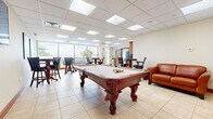 Located at 330 W First St in Dayton, Ohio, this single-floor community lounge and recreation room offers a versatile, open-plan space designed for social gathering and leisure. The room is finished with neutral large-format ceramic tile flooring and a drop ceiling with recessed panel lighting that keeps the interior bright and welcoming throughout.

At the heart of the space sits an ornately carved hardwood pool table with tassel pockets, complemented by a wall-mounted flat-screen television and a leather sofa, creating a dedicated game and entertainment zone. A structural column divides the room subtly, adding architectural interest while preserving an open feel.

Multiple bar-height round tables paired with upholstered swivel chairs provide ample casual seating throughout. Colorful abstract artwork adorns the walls, adding a lively, contemporary touch to the otherwise neutral palette.

A kitchenette area along one wall features warm wood cabinetry, a full-size refrigerator, and a sink, providing convenient service for gatherings. Large floor-to-ceiling windows along one side of the room flood the interior with natural light and offer direct views of the outdoor pool deck and terrace, where lounge chairs and umbrellas are visible. Potted plants positioned near the windows enhance the connection to the outdoors, contributing to an inviting, relaxed atmosphere throughout the space.