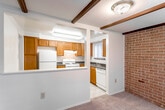This single-floor residence offers a thoughtfully designed layout that balances comfort and functionality throughout its living spaces. The kitchen serves as a practical centerpiece, featuring warm honey-toned wood cabinetry, dark countertops, and a full suite of white appliances including a gas range, refrigerator, and dishwasher. A window above the sink brings in natural light, while a pass-through peninsula connects the kitchen to the adjacent living area. Distinctive decorative wood beams accent the ceiling at the kitchen-living area transition, adding architectural character to the space. A full-height exposed brick wall anchors the living area, providing a bold and textural focal point that lends warmth and character to the room. The living area is generously sized, featuring neutral carpet and large windows that allow ample natural light to fill the space. The bedrooms are comfortably proportioned with plush carpeting, neutral walls, and ample closet space, including a walk-in closet in the primary bedroom. The bathroom is clean and bright, featuring a full soaking tub with tiled surround and a window overlooking lush greenery. Overall, the space presents a cozy and classic aesthetic with durable, functional finishes throughout.