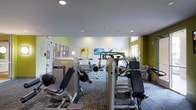This fitness center offers a well-rounded exercise environment designed to support a variety of workout routines. The space is outfitted with a diverse array of equipment, including multiple strength-training machines such as leg press and cable resistance units, as well as cardio equipment including stationary bikes and rowing machines. A wall-mounted flat-screen television is positioned at the front of the room, providing entertainment during workouts. The flooring features a patterned, multi-tonal carpet designed to withstand heavy equipment use, while recessed lighting in the white ceiling ensures the space is thoroughly and evenly illuminated. The walls are accented with a vibrant yellow-green tone, adding an energetic ambiance to the room. Decorative artwork depicting nature scenes enhances the visual appeal of the space. Large windows along the perimeter allow natural light to filter in, contributing to an open and airy atmosphere. The single-floor layout maximizes accessibility, with equipment arranged to allow comfortable movement throughout the space. Overall, the fitness center is thoughtfully designed to provide users with a functional, motivating, and well-lit environment for achieving their health and wellness goals.