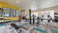This modern fitness center is thoughtfully designed to support a comprehensive workout experience across multiple distinct zones. The main exercise floor features patterned carpet tiles accented with teal highlights, lending the space a contemporary, energetic aesthetic. Recessed lighting and ceiling fans keep the environment comfortable and well-lit throughout.

The strength training area is outfitted with a robust selection of selectorized resistance machines, cable stations, a dumbbell rack, and adjustable benches, catering to a full range of muscle groups. Large windows lining the perimeter flood the space with natural light and offer views of the surrounding community.

Wall-mounted mirrors expand the sense of space and allow users to monitor their form. A vibrant yellow accent wall, adorned with a sweeping, colorful wave mural, adds a bold artistic touch and energizes the environment. A hallway connects the main gym floor to additional areas, maintaining an open and navigable layout.

A separate studio room with light hardwood flooring provides a dedicated area for functional and flexibility training, stocked with stability balls, kettlebells, resistance bands, and a stationary bike. Floor-to-ceiling windows in this room maximize natural light. An additional room with hardwood flooring and a glass-paneled door offers a more private space for cycling or individual workouts.