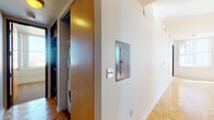 This single-floor residence presents a thoughtfully designed layout that balances functionality and urban living. The main living area is a bright, inviting space finished with warm parquet hardwood flooring that reflects abundant natural light pouring in through multiple large windows. These windows frame expansive city skyline views, reinforcing the elevated urban character of the home.

The kitchen is defined by light maple cabinetry paired with dark countertops, stainless steel appliances—including a refrigerator, range, and over-the-range microwave—and a peninsula that separates the cooking area from the living space while providing additional prep and seating potential. Parquet flooring continues seamlessly throughout.

The bedroom is carpeted for comfort and features two sets of sliding wood-panel closet doors, offering generous storage. A large window fills the room with natural light and city views.

Two bathrooms serve the residence: one equipped with a tiled walk-in shower and a wood-toned vanity with dark countertop, and a second full bathroom featuring a tub-shower combination and matching vanity with a large mirror.

A hallway utility closet houses a stacked washer and dryer unit, adding everyday convenience. An electrical panel is also accessible along the corridor, which features the same parquet flooring found throughout the main living spaces.