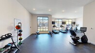 This fitness center in Memphis, TN offers a thoughtfully designed, single-floor exercise space that provides a comprehensive workout environment. The room features a clean, modern aesthetic with crisp white walls, recessed lighting, and durable dark rubber flooring throughout, creating a functional and inviting atmosphere.

Large black-framed windows line multiple walls, flooding the space with abundant natural light and offering views of the surrounding outdoor area. Wall-mounted flat-screen televisions are positioned near the cardio equipment for entertainment during workouts.

The cardio section is well-appointed with multiple treadmills, elliptical trainers, and a stationary recumbent bike, all arranged to take full advantage of the natural light from the windows. A dedicated free weights area includes a tiered dumbbell rack, kettlebell rack, stability balls, and weight benches, supporting a variety of strength and functional training routines. A medicine ball rack with color-coded balls rounds out the strength training offerings.

The space also features an open floor area suitable for stretching or bodyweight exercises, along with a step platform for aerobic training. The layout is efficient and uncluttered, allowing for comfortable movement between equipment zones. The entry area includes a glass-panel door and mirror, adding to the open, airy feel of this well-maintained facility.