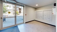 This building lobby serves as the central hub for residents and visitors, offering a clean and functional layout with modern design elements. The main entrance features full-height glass double doors that allow natural light to flood the entryway, providing clear sightlines to the exterior. A durable tile floor in a neutral tone extends throughout the lobby area, complemented by dark baseboards that add a subtle contrast to the light-colored walls.

A prominent bank of individual mailboxes is mounted along the wall near the entrance, providing convenient mail access for all building residents. Adjacent to the mailbox area, a passenger elevator with clearly marked call buttons and safety certifications offers vertical access to the upper floors. Recessed and flush-mounted circular ceiling fixtures provide consistent, even illumination throughout the space.

From the lobby, a corridor extends further into the building, transitioning from tile flooring to a patterned, textured carpet in earthy tones. The hallway is lined with neutral walls, dark baseboards, and evenly spaced overhead lights, leading residents efficiently to their units. Emergency signage, a fire alarm system, and an intercom panel are thoughtfully integrated throughout, ensuring safety and accessibility. The overall ambiance is clean, contemporary, and practical.