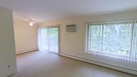 This single-floor space offers a bright and open living area designed for comfort and functionality. The main living space is finished with neutral-toned carpet and light-colored walls, creating a clean and airy atmosphere. Two large windows fitted with vertical blinds allow generous amounts of natural light to filter in, while framing views of surrounding greenery that lend the interior a serene, nature-connected feel. A sliding glass door provides direct access to an outdoor balcony, further enhancing the connection to the outdoors and offering an additional area for relaxation. Climate control is addressed through a wall-mounted air conditioning unit, while baseboard heating units run along the perimeter of the room, ensuring year-round comfort. A ceiling fan is also present, contributing to air circulation throughout the space. The overall layout is open and unobstructed, providing flexibility for various furniture arrangements. The combination of natural light, outdoor access, and green views makes this a pleasant and functional living environment.
