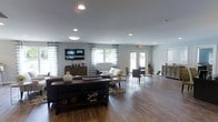This single-floor space presents an open, airy layout designed for comfort and functionality. The main gathering area features rich hardwood-style flooring that runs throughout, lending warmth and a cohesive feel to the entire space. Recessed lighting is embedded throughout the smooth, light-toned ceiling, complementing the generous natural light that pours in through multiple windows dressed with elegant patterned curtains and French doors at the rear.

The living area is thoughtfully arranged with a large, dark wood console table as a focal point, accompanied by a mounted flat-screen television, making it ideal for group entertainment or casual gatherings. A comfortable sofa and coordinating accent chairs are arranged to encourage conversation. Adjacent to this seating area, a bistro-style dining arrangement features darker chairs and small round side tables, offering a relaxed space for small group interaction.

Toward the rear, French doors open to what appears to be an outdoor area, further enhancing the sense of openness and connectivity to the outdoors. A secondary counter or service area is visible along the far wall, adding a practical utility element to the space. Overall, the design strikes a balance between modern aesthetics and inviting functionality, making it well-suited as a communal lounge or meeting area.
