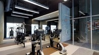 This single-floor fitness center is thoughtfully designed to provide a well-equipped and visually dynamic workout environment. The space features a sleek, contemporary aesthetic defined by dark-painted walls and ceilings that create a focused, high-energy atmosphere. Linear pendant lighting runs overhead, supplemented by recessed ceiling lights, ensuring the entire floor is evenly and effectively illuminated.

A standout architectural detail is the textured stone column near the entrance, which adds a bold visual contrast against the darker surroundings and contributes to the space's industrial-modern character. Floor-to-ceiling glass panels along one side allow natural light to filter in and provide a sense of openness and connectivity to adjacent areas.

The gym is outfitted with a comprehensive selection of Matrix brand equipment, including stationary bikes, cable machines, and strength-training stations, arranged to allow comfortable circulation throughout the floor. Large wall mirrors extend the visual depth of the space while enabling users to monitor their form. The flooring transitions between carpet and rubberized mat zones, delineating cardio and strength training areas. Overall, the space balances form and function effectively.