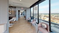 This upscale amenity floor is designed to impress, combining contemporary aesthetics with an array of social and recreational spaces set high above the city skyline. Floor-to-ceiling glass walls run along the perimeter of the lounge area, flooding the interior with natural light and offering sweeping panoramic views of the surrounding urban landscape. Light wood flooring anchors the open-plan lounge, which features a mix of plush seating arrangements — from rounded accent chairs in soft blush tones to a generous leather sectional sofa — all oriented to take in the remarkable cityscape. Large sliding glass doors open seamlessly onto a spacious outdoor terrace, extending the living area into the open air and providing an inviting setting for gatherings against a dramatic skyline backdrop. A wide corridor connects the lounge to the rest of the floor, maintaining the same airy feel with continued floor-to-ceiling glazing on one side. The game room offers a dynamic contrast, featuring a pool table, foosball table, wall-mounted flat-screen televisions, and a bar-height seating area set against richly textured dark accent walls with subtle LED underlighting. Checkered carpet tiles add visual interest underfoot, while recessed lighting and corner windows with city views complete the polished, energetic atmosphere of this exceptional amenity space.