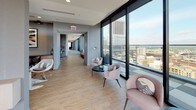 This upscale amenity floor is designed to impress, combining contemporary aesthetics with an array of social and recreational spaces set high above the city skyline. Floor-to-ceiling glass walls run along the perimeter of the lounge area, flooding the interior with natural light and offering sweeping panoramic views of the surrounding urban landscape. Light wood flooring anchors the open-plan lounge, which features a mix of plush seating arrangements — from rounded accent chairs in soft blush tones to a generous leather sectional sofa — all oriented to take in the remarkable cityscape. Large sliding glass doors open seamlessly onto a spacious outdoor terrace, extending the living area into the open air and providing an inviting setting for gatherings against a dramatic skyline backdrop. A wide corridor connects the lounge to the rest of the floor, maintaining the same airy feel with continued floor-to-ceiling glazing on one side. The game room offers a dynamic contrast, featuring a pool table, foosball table, wall-mounted flat-screen televisions, and a bar-height seating area set against richly textured dark accent walls with subtle LED underlighting. Checkered carpet tiles add visual interest underfoot, while recessed lighting and corner windows with city views complete the polished, energetic atmosphere of this exceptional amenity space.