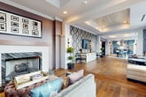 This elegantly designed single-floor space blends classical architectural details with bold, contemporary accents to create a refined and welcoming environment. The entry corridor sets a polished tone with herringbone hardwood flooring, tall wainscoting, ornate panel molding, and amber glass chandeliers overhead. A decorative medallion wall art piece anchors the reception area at the corridor's end.

The lounge area features soaring double-height ceilings and expansive floor-to-ceiling windows that flood the space with natural light and offer views of surrounding greenery and the urban skyline. Patterned encaustic-style cement tile floors complement a curated mix of furnishings, including a deep navy tufted Chesterfield sofa, sculptural velvet accent chairs, and an ornately carved coffee table.

Adjacent to the lounge, a refreshment area makes a dramatic statement with floor-to-ceiling white marble-veined wall cladding and a striking suspended installation of colorful glass bottles. Powder-blue shaker-style cabinetry with brass hardware lines the walls beneath a crisp white countertop, paired with bistro-style seating. The overall ambiance throughout is luxurious yet approachable, combining classical craftsmanship with artful, playful design elements.