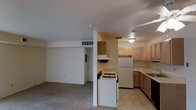 This single-floor space presents an open-concept design that connects the kitchen and living area through a partial dividing column. The kitchen is equipped with essential appliances, including a white gas range with an overhead range hood, a full-size refrigerator, and a double-basin sink set within laminate countertops. Light wood cabinetry lines the walls, providing ample storage, while tile flooring throughout the kitchen offers easy maintenance. A ceiling-mounted light fixture provides overhead illumination. The adjacent living area features wall-to-wall carpeting and a neutral color palette, creating a comfortable and understated atmosphere. A central HVAC vent is positioned along the upper wall, ensuring climate control throughout the space. The layout benefits from a straightforward flow between functional zones, with a doorway visible at the rear offering access to additional areas of the unit. Flush ceilings and simple finishes characterize the overall aesthetic, making the space well-suited for practical, everyday living.