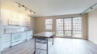 This single-floor studio with one bathroom offers an open, flexible layout enhanced by wood-style flooring that flows consistently throughout the space. The main living area is bright and airy, anchored by a large bank of floor-to-ceiling windows that frame views of the surrounding urban skyline, filling the interior with generous natural light.

The kitchen is positioned along one wall and is well-equipped with white cabinetry — including upper glass-front cabinets and open shelving — granite countertops, a stainless steel sink with a modern pull-down faucet, and a full suite of white appliances including a refrigerator, electric range, dishwasher, and built-in microwave. Track lighting overhead provides focused illumination across the workspace.

A barn-style sliding door leads to a dedicated sleeping area, offering a degree of separation within the open floor plan. Adjacent to this space is a walk-in closet with built-in shelving and hanging rods, providing ample storage.

The bathroom features a granite-topped vanity with white cabinetry, a tub-shower combination with white tile surround, and a patterned tile floor. A small window introduces natural light into the bathroom as well. Overall, the studio presents a clean, contemporary aesthetic well-suited to efficient urban living.
