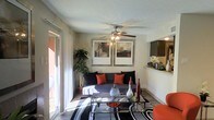 This single-floor residence centers around a thoughtfully designed living area that blends comfort and contemporary style. The living room is highlighted by a ceiling fan with integrated lighting, providing both air circulation and ambient illumination. A textured ceiling adds subtle depth to the space overhead.

Natural light flows generously into the room through large sliding glass doors fitted with sheer white curtains, offering a view of and direct access to an outdoor area. The neutral beige walls serve as a versatile backdrop, complemented by a pair of framed wall art pieces that add a refined visual focal point above the seating area.

The living space is furnished with a gray sofa accented by bold orange and black throw pillows, anchored by a glass-top coffee table resting on a black-and-white patterned area rug. Decorative vases and accessories on the coffee table contribute to the curated aesthetic. A tall indoor plant adds a natural, organic element to the corner of the room.

A pass-through opening on one side of the living area connects to an adjacent kitchen or bar space, enhancing the open, communicative layout of the home and supporting easy interaction between living and functional areas.