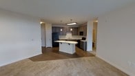 This single-floor residence offers a thoughtfully designed open-concept layout that seamlessly connects the kitchen and living areas, creating a spacious and functional environment. The kitchen is a standout feature, showcasing dark taupe cabinetry that extends to the upper walls, paired with a clean white countertop and a matching peninsula island with seating overhang — ideal for casual dining or entertaining. Black appliances, including a full-size refrigerator, dishwasher, and range, provide a striking contrast against the light cabinetry and white backsplash. Pendant lighting hangs above both the island and the main prep area, adding a modern, polished touch. The transition from the kitchen's hard flooring to the plush carpeted living area creates a natural delineation between spaces while maintaining an open flow. Neutral tones throughout — from the soft carpet to the crisp white walls — create a bright and welcoming ambiance. A doorway visible beyond the kitchen hints at additional rooms, including what appears to be a bathroom or hallway, further extending the functional layout of this well-appointed residence.