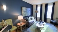 This single-floor space makes a striking first impression with its bold design choices and carefully curated interiors. The living area serves as the centerpiece, anchored by a deep navy blue accent wall that sets a dramatic yet refined tone throughout. A large-scale abstract painting adorns the feature wall, adding an artistic focal point that draws the eye and elevates the room's sophistication.

The living space is thoughtfully furnished with a plush gray sofa accented by an array of decorative pillows in complementary navy and sage green tones. A round white coffee table sits atop a richly patterned area rug that introduces warm greens and blues, tying the color palette together seamlessly. A rustic wooden side table paired with a decorative table lamp adds warmth and contrast to the cooler tones of the room.

Natural light flows in generously through windows dressed with floor-length navy curtains, which can be drawn to create an intimate atmosphere or opened to brighten the space. A modern arc floor lamp provides additional ambient lighting, enhancing the layered lighting design.

The overall ambiance balances boldness with comfort — a space that feels both design-forward and livable, ideal for relaxed entertaining or quiet everyday living.