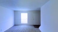 This two-floor space presents a clean and straightforward layout with a neutral color palette throughout. The pictured room features light gray carpeting that extends across the floor, complemented by white walls and a textured ceiling that adds subtle visual interest overhead. A paneled white door serves as a focal point along the far wall, flanked by a window fitted with vertical blinds that allow natural light to filter into the room, creating a bright and open atmosphere. The transition near the doorway reveals a change to dark hardwood or laminate flooring, suggesting a distinct entry or adjoining area with a different finish. The room's simple, uncluttered design provides a versatile foundation adaptable to a variety of uses. Spanning two floors, the overall space offers functional separation between living areas, with the layout providing flexibility for different room configurations. The consistent use of white tones across walls, ceilings, and doors gives the interior a cohesive and airy quality throughout.