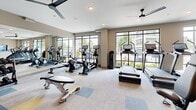 Located at 500 Congaree Rd in Greenville, SC, this well-appointed fitness center spans a single floor and is thoughtfully divided into two distinct workout zones. The main cardio and strength training area features neutral-toned carpet with colorful accent tiles, floor-to-ceiling grid-frame windows that flood the space with natural light and offer views of the surrounding streetscape. This zone is equipped with multiple treadmills, ellipticals, stationary bikes, a cable machine, free weights, and a wall-mounted mirror that enhances the sense of space. Ceiling fans provide added comfort throughout. The second zone serves as a dedicated studio or functional training area, finished with warm hardwood flooring, soft blue-gray walls, and a tray ceiling. This room houses spin bikes, a large wall-mounted display screen, and a comprehensive wall-mounted equipment rack stocked with kettlebells, medicine balls, resistance bands, TRX suspension trainers, and dumbbells. A heavy-duty tire sled adds a functional fitness element. Double glass-pane doors with black framing connect the two zones, creating an open, cohesive feel. The overall design is clean, modern, and well-lit, offering a comprehensive fitness experience.