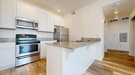 This single-floor residence is designed with a clean, contemporary aesthetic that emphasizes bright, open spaces throughout. The kitchen serves as a central highlight, showcasing white shaker-style cabinetry extending to the ceiling for generous storage, speckled granite countertops, and a suite of stainless steel appliances including a range, over-the-range microwave, and refrigerator. A peninsula with matching granite provides additional workspace and casual seating, functioning as a natural divider between the kitchen and the adjoining living area.

The living area features warm wood-style plank flooring that runs throughout the main spaces, complemented by white walls and recessed lighting that enhance the sense of brightness. A large window allows natural light to pour in, offering views of the surrounding area and creating an airy atmosphere.

The bedrooms are finished with plush neutral carpet and feature multiple windows fitted with horizontal blinds, allowing for controlled natural light. The bathroom is outfitted with a full soaking tub and shower combination, a white vanity with an integrated sink, and warm wood-tone plank flooring. The overall palette of whites and neutrals throughout the unit creates a cohesive, modern living environment.