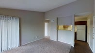 This single-floor residential space is designed with a practical and open layout that maximizes functionality. The living area is spacious and welcoming, featuring wall-to-wall carpeting in a warm neutral tone that adds comfort underfoot. The walls are finished in a soft gray palette, creating a calm and versatile backdrop suited to a variety of interior styles. A textured ceiling completes the classic look of the main living space.

A standout feature of this space is the pass-through kitchen bar, which connects the kitchen to the living area, offering a convenient and social arrangement ideal for everyday living. The kitchen is partially visible through the pass-through opening, revealing warm wood-toned cabinetry and under-cabinet lighting that adds a welcoming glow. HVAC vents are integrated into the ceiling, ensuring year-round climate comfort.

A hallway leads from the living area toward additional rooms, suggesting a thoughtful floor plan that separates common and private spaces. The overall ambiance of the space is clean, neutral, and move-in ready, offering a solid foundation for personalization. The combination of open sightlines, functional kitchen access, and consistent neutral finishes throughout makes this a practical and comfortable single-floor residence.