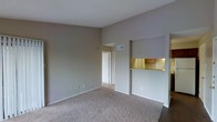 This single-floor residential space is designed with a practical and open layout that maximizes functionality. The living area is spacious and welcoming, featuring wall-to-wall carpeting in a warm neutral tone that adds comfort underfoot. The walls are finished in a soft gray palette, creating a calm and versatile backdrop suited to a variety of interior styles. A textured ceiling completes the classic look of the main living space.

A standout feature of this space is the pass-through kitchen bar, which connects the kitchen to the living area, offering a convenient and social arrangement ideal for everyday living. The kitchen is partially visible through the pass-through opening, revealing warm wood-toned cabinetry and under-cabinet lighting that adds a welcoming glow. HVAC vents are integrated into the ceiling, ensuring year-round climate comfort.

A hallway leads from the living area toward additional rooms, suggesting a thoughtful floor plan that separates common and private spaces. The overall ambiance of the space is clean, neutral, and move-in ready, offering a solid foundation for personalization. The combination of open sightlines, functional kitchen access, and consistent neutral finishes throughout makes this a practical and comfortable single-floor residence.