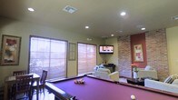 This single-story space is designed as a multifunctional entertainment and lounge area. The room's most prominent feature is a full-sized pool table with a rich purple felt surface, positioned centrally to allow ample room for play. Recessed lighting in the ceiling provides even illumination throughout, complementing the natural light that filters in through multiple large windows fitted with wooden horizontal blinds. The windows offer views of an outdoor setting and contribute to the bright, open atmosphere of the room.

A notable architectural detail is the exposed brick accent wall, which adds texture and a warm, rustic character to the space, contrasting effectively with the smooth, light-colored walls elsewhere in the room. A wall-mounted flat-screen television is positioned in the corner, making it easily visible from multiple vantage points. Adjacent to the TV, a comfortable lounge area features upholstered seating, creating a relaxed gathering spot. Decorative elements, including framed wall art and floral arrangements, add personality and warmth. The overall ambiance balances a casual, social atmosphere with practical entertainment amenities.