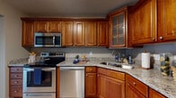 This single-floor residence offers a thoughtfully designed layout that balances functionality and comfort throughout its well-maintained spaces. The kitchen serves as a standout feature, showcasing rich, warm-toned wood cabinetry with raised-panel doors and glass-front accent cabinets. Granite countertops in a speckled neutral palette provide ample workspace, while a full suite of stainless steel appliances — including an electric range, built-in microwave, dishwasher, and double-basin sink — delivers a complete cooking experience. Light tile flooring grounds the kitchen and transitions naturally into the adjoining hallway and entry areas. A half-wall pass-through with a granite ledge connects the kitchen to the main living area, fostering an open and communicative flow between spaces. The living area is generously proportioned, featuring soft wall-to-wall carpeting, neutral taupe walls, and textured ceilings. Natural light filters in through windows visible along the perimeter. The bathroom includes a full soaking tub and shower combination set against beige tile surround. A dedicated in-unit laundry closet houses a stacked washer and dryer, adding everyday convenience. Closet storage and carpeted bedroom spaces complete this practical, move-in-ready home.