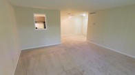 This single-floor residential space offers a clean, functional layout designed for comfortable everyday living. The main living area is generously proportioned, featuring plush wall-to-wall carpeting in a neutral tone that lends warmth and softness underfoot. Light-colored walls throughout create a bright, airy atmosphere, and a ceiling fan positioned centrally provides both ventilation and overhead lighting for the space.

A distinctive architectural detail is the pass-through window cutout that connects the living area to the kitchen, offering both visual openness and practical convenience. The kitchen, visible through the pass-through, features cabinetry in a warm wood tone and includes standard appliances, making efficient use of the available space.

The entryway is neatly defined, with a white paneled door that provides a welcoming transition into the unit. HVAC ventilation is integrated into the upper wall, ensuring year-round climate comfort. The overall design palette — soft neutrals, white trim, and warm wood accents — creates a cohesive and inviting ambiance. The space is well-suited for those seeking a straightforward, move-in-ready layout with thoughtful connectivity between living and cooking areas.