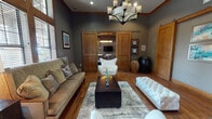 This single-floor space exudes a refined, lodge-inspired aesthetic defined by a cohesive palette of deep charcoal gray walls, warm hardwood floors, and natural wood accents throughout. The main living area serves as the heart of the space, anchored by a statement chandelier with candle-style lighting, an area rug, and a comfortable seating arrangement featuring a neutral-toned sofa, tufted white accent chairs, and a dark wood chest coffee table. A standout architectural detail is the set of sliding barn-style doors crafted from rich wood, which open to reveal an adjoining media room equipped with a wall-mounted television. Tall, wood-framed windows with horizontal blinds allow generous natural light to filter in while offering views of the surrounding greenery. The vaulted ceiling height in the window area adds an impressive sense of verticality to the living space. An additional private room, separated by a wood-paneled door with frosted glass, features carpet flooring and a classic mahogany chest, offering a quieter, more intimate setting. The space connects further to a brighter adjoining area with lighter walls and tile flooring, providing a natural contrast to the moody, sophisticated tones of the main rooms. Throughout, wood trim and molding details reinforce the space's warm, craft-forward character.