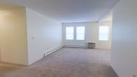This single-floor residential space offers a clean, straightforward layout designed for comfortable everyday living. The main living area is notably open and well-proportioned, featuring neutral wall-to-wall carpeting that lends a warm, understated feel to the interior. Crisp white walls provide a blank canvas, making the space highly adaptable to a variety of furnishing styles and personal tastes.

Natural light is a defining characteristic of this space, with multiple windows allowing daylight to filter in and brighten the interior. The windows are fitted with blinds, offering adjustable light control and privacy as needed. Baseboard heating units run along the perimeter of the living area, ensuring efficient and consistent warmth throughout the colder months. An additional wall-mounted HVAC unit provides supplemental climate control, offering flexibility for year-round comfort.

The overall ambiance is calm and neutral, with a minimalist aesthetic that emphasizes functionality and ease of use. The single-floor layout ensures convenient, step-free access to all areas of the space. Whether used as a primary residence or transitional living space, this unit provides a solid, practical foundation with essential amenities already in place.