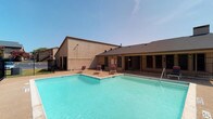 Located in Sherman, this outdoor amenity space centers around a generously sized swimming pool with clearly marked depth indicators, offering both shallow and deeper sections to accommodate a range of swimmers. The pool is surrounded by a broad concrete deck furnished with red-and-white lounge chairs, providing ample space for sunbathing and relaxation under the open Texas sky. A low-rise clubhouse building adjoins the pool area, featuring a covered, gated patio that offers shaded seating and views of the surrounding grounds. The clubhouse is constructed with a combination of brick and stucco exterior finishes beneath a pitched, shingled roof, and includes interior-access doors visible through the iron security fencing. The entire pool area is enclosed by a black wrought-iron fence, ensuring safety and security. The surrounding residential buildings are constructed of classic brick and feature balconies, adding a sense of community to the setting. Mature trees dotting the perimeter provide a natural backdrop and some relief from the sun. The overall atmosphere is open and inviting, with the bright, clear water of the pool serving as the focal point of this well-maintained outdoor community space.