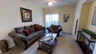 This single-floor residence offers a comfortable and well-appointed living space designed for everyday functionality and relaxed living. The living room serves as the heart of the home, featuring plush, dark-toned upholstered seating — including a generously sized sofa and a coordinating armchair — arranged around a glass-top coffee table with ornate wood detailing. Soft, neutral-toned carpeting runs throughout, adding warmth underfoot and contributing to the room's inviting atmosphere. A ceiling fan with an integrated light fixture is centrally positioned overhead, providing both air circulation and ambient lighting. Natural light pours into the space through a sliding glass door fitted with vertical blinds, offering a view of lush greenery outside and providing convenient access to the outdoors. The walls are finished in a soft, warm neutral tone and are accented with framed decorative artwork, lending a polished, lived-in character to the space. The overall ambiance is cozy and welcoming, with a traditional design aesthetic that balances comfort and style throughout.