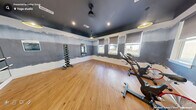 This dedicated fitness studio is thoughtfully designed to maximize function and atmosphere within a compact footprint. The space is defined by a striking dark-painted ceiling that creates a bold, energizing aesthetic, contrasted by white wainscoting along the lower walls and a distinctive gradient painted mural in muted blue-grey tones spanning the upper wall surfaces.

Natural light flows in through multiple roller shade-equipped windows along two walls, offering outdoor views while maintaining adjustable light control. Warm-toned hardwood-style flooring runs throughout, providing a durable and visually appealing workout surface.

The studio is equipped with several spin bikes positioned for group cycling sessions, a wall-mounted flat-screen television, and a full dumbbell rack with a range of weights. A large mirrored wall with a ballet barre attachment enhances the sense of space and supports a variety of workout styles. Step platforms and additional small equipment are also present.

Pendant lighting with gold-accented shades complements the recessed ceiling lights, creating a layered and inviting ambiance. A glass-panel door with a dark frame provides access to an adjoining space, completing this well-designed, multipurpose fitness environment.