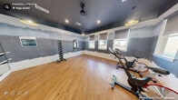 This dedicated fitness studio is thoughtfully designed to maximize function and atmosphere within a compact footprint. The space is defined by a striking dark-painted ceiling that creates a bold, energizing aesthetic, contrasted by white wainscoting along the lower walls and a distinctive gradient painted mural in muted blue-grey tones spanning the upper wall surfaces.

Natural light flows in through multiple roller shade-equipped windows along two walls, offering outdoor views while maintaining adjustable light control. Warm-toned hardwood-style flooring runs throughout, providing a durable and visually appealing workout surface.

The studio is equipped with several spin bikes positioned for group cycling sessions, a wall-mounted flat-screen television, and a full dumbbell rack with a range of weights. A large mirrored wall with a ballet barre attachment enhances the sense of space and supports a variety of workout styles. Step platforms and additional small equipment are also present.

Pendant lighting with gold-accented shades complements the recessed ceiling lights, creating a layered and inviting ambiance. A glass-panel door with a dark frame provides access to an adjoining space, completing this well-designed, multipurpose fitness environment.