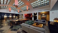 This striking hotel lobby is defined by its standout architectural centerpiece: a sweeping geometric skylight ceiling crafted from a latticed grid of glass panels, suspended pendant lanterns casting warm amber light across the open lounge below. The reception area greets guests with a sleek, dark-paneled front desk set against high white walls and a bold dark ceiling, creating a sophisticated contrast. Polished concrete and terrazzo flooring with diamond-patterned inlays extend throughout the space, adding a vintage character that complements the eclectic design.

The expansive lounge is thoughtfully arranged with clusters of sofas, armchairs, and a long communal dining table, offering guests both intimate and social settings. A bar — visible through a darkened doorway — adds a lively focal point. Along the corridors, gallery-style walls display an eclectic mix of framed photographs, maps, artwork, and directional signage, contributing to an atmosphere of curated charm. Leather armchairs and warm table lamps are positioned throughout, inviting guests to linger.

The elevator foyer is a moody, dramatic contrast, featuring deep navy and near-black walls, metallic elevator doors, geometric terrazzo floors, and lush potted plants that add organic warmth to the bold, dark palette.