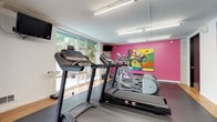 This single-floor amenity space is thoughtfully designed to support both fitness and productivity. The fitness area is well-appointed with multiple treadmills, elliptical trainers, a stationary bike, and a multi-station weight machine, all arranged on protective rubber flooring. Large windows flood the workout space with natural light and offer views of lush greenery outdoors, creating an energizing atmosphere. Overhead fluorescent lighting ensures consistent illumination throughout. A vibrant pink accent wall featuring a bold, colorful work of art adds a dynamic visual element to the exercise environment. A dumbbell rack and additional free weights are conveniently positioned along a hardwood-floored corridor, which connects the various areas via sleek glass partition doors. Adjacent to the fitness space, a compact business center provides two computer workstations set along a continuous white desk, paired with ergonomic orange chairs. Wall-mounted open shelving with decorative accents lines the charcoal accent wall above the desks, while soft recessed lighting creates a focused, professional environment. The overall design balances functionality with contemporary aesthetics, offering users a cohesive and versatile facility suited for both physical activity and work.