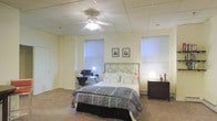 This single-floor space centers around a generously sized bedroom that combines comfort and functionality. The room features a dropped acoustic tile ceiling equipped with a ceiling fan and light fixture, providing both air circulation and ambient lighting. Soft, wall-to-wall carpeting runs throughout, adding warmth underfoot.

The bedroom is anchored by a full-sized bed with a decorative metal headboard featuring circular geometric details, dressed in a classic patterned comforter. A dark-stained wooden dresser sits to one side, topped with a contemporary globe lamp and decorative accents. Framed artwork adorns the light-painted walls, contributing a personal and curated touch to the space.

Natural light filters in through multiple windows fitted with horizontal blinds, allowing for adjustable privacy and brightness. A dedicated workspace is set up in one corner, complete with a desk, computer monitor, and a task lamp, making the room well-suited for both rest and productivity. A baseboard heating unit is also visible, ensuring year-round comfort.

The overall ambiance is calm and functional, with a neutral color palette of soft yellows and warm beiges that creates an inviting and livable atmosphere throughout the space.