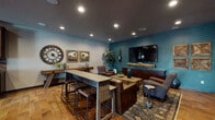 Located at 16511 East Sprague Avenue in Spokane, WA, this single-floor community clubhouse blends rustic warmth with modern amenities throughout its thoughtfully designed spaces. Rich wood-look tile and hardwood flooring flow seamlessly across the entire facility, anchoring a cohesive and inviting aesthetic.

The central lounge greets visitors with soaring ceilings, a statement chandelier, and a grouping of leather wingback chairs arranged around a round coffee table atop a patterned area rug. A dark-tile-surround fireplace with built-in shelving adds a cozy focal point, while large windows and glass entry doors allow generous natural light to fill the room.

Adjacent to the lounge, a fully equipped kitchen features dark cabinetry, granite countertops, stainless steel appliances, and a bar-height island with seating — ideal for community gatherings and events. Sliding barn doors crafted from rich wood add a rustic-chic architectural detail that connects this area to the broader space.

A dedicated media lounge offers comfortable leather seating, a large wall-mounted flat-screen TV, and a high-top work table, all set against a deep teal accent wall. High clerestory windows in the casual seating nooks provide additional natural light and exterior views.

Rounding out the facility is a well-equipped fitness center visible in the floor plan, completing this versatile and stylishly appointed community hub.