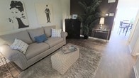 This thoughtfully designed two-floor space combines modern sophistication with comfortable, curated interiors. The living area immediately sets the tone with its striking contrast of light and dark — a bold, dark accent wall anchors the room while neutral tones in the furnishings create a balanced and inviting atmosphere. Rich hardwood flooring runs throughout, adding warmth and continuity to the space.

The living room is appointed with a generously sized, taupe upholstered sofa adorned with nailhead trim detail and complementary blue accent pillows, evoking a sense of relaxed elegance. A patterned ottoman serves as a functional centerpiece atop a plush, textured area rug. Artistic touches are present throughout, including a bold monochromatic wall print that adds a classic, eclectic character to the room.

A sleek black storage cabinet and a gold-toned console table provide both style and practicality, while a tall indoor palm plant introduces a natural, organic element to the interior. Subtle, ambient lighting from a table lamp enhances the warm, intimate feel of the space.

Spanning two floors, this property offers a well-considered layout that balances open, communal living areas with the privacy and functionality expected across multiple levels — making it a versatile and visually compelling space.