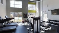 Located at 811 Spring Forest Rd in Raleigh, NC, this fitness center is designed to support an active lifestyle within a vibrant residential community. The space features impressively high ceilings with exposed ductwork, lending an industrial-modern aesthetic that feels open and energizing. Large, multi-pane windows line the walls, flooding the interior with abundant natural light while offering direct views of the outdoor pool area and surrounding community buildings. Accent walls in bold chartreuse yellow add a dynamic visual contrast against the crisp white walls, contributing to an upbeat, motivating atmosphere. The facility is equipped with a range of Life Fitness cardio and strength-training machines, including treadmills, ellipticals, and cable resistance equipment, all arranged on dark rubberized flooring designed for durability and safety. The layout is thoughtfully organized to allow comfortable circulation between stations. The combination of natural light, modern equipment, and contemporary design elements creates a welcoming and functional environment well-suited for daily workouts.