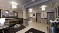 This sophisticated lobby presents a refined first impression with its carefully considered design and upscale finishes. The space is anchored by a neutral beige and cream color palette, accented by a bold black wainscoting band that runs along the lower walls, creating a striking visual contrast throughout. Structural columns with detailed millwork add architectural character, while ornate crystal-accented pendant and chandelier-style light fixtures cast a warm, inviting glow across the tiled floors. A decorative monogram emblem — prominently displayed on the wall near the elevator bank — serves as a distinguished focal point and brand identifier for the building. The elevator area is neatly integrated into the design, finished with brushed metallic doors and complementary wall paneling. A comfortable seating area with dark upholstered furniture and a textured ottoman rests atop a patterned area rug, offering a welcoming space for residents or guests. A large framed mirror enhances the sense of openness, while potted greenery adds a touch of natural warmth. Staircase access is also visible, reinforcing the multi-functional nature of this well-appointed entrance space.