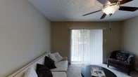 This single-floor living space centers around a comfortable living room designed with a neutral, understated aesthetic. The room features warm taupe-toned walls that create a relaxed atmosphere, complemented by wall-to-wall carpeting that adds softness underfoot. A textured popcorn ceiling adds subtle dimension to the space, while a ceiling fan with an integrated light fixture provides both air circulation and overhead lighting.

A prominent sliding glass door serves as the focal point of the living room, fitted with vertical blinds that allow for adjustable natural light and privacy control. The door provides a connection to an outdoor area, allowing natural light to filter into the room during the day.

The living area accommodates a white upholstered sofa accented with dark decorative pillows, paired with a sleek, rounded coffee table, illustrating the room's functional layout. The overall ambiance of the space is calm and practical, with simple finishes and a straightforward design that offers a comfortable, livable environment. The single-floor layout ensures easy accessibility throughout the unit.