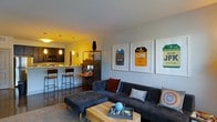 This single-floor residence offers a thoughtfully designed open-concept layout that blends living and kitchen spaces into one cohesive, functional area. The living room is anchored by a large, dark sectional sofa accented with colorful throw pillows and paired with a tufted ottoman and a natural wood coffee table, creating a comfortable yet contemporary gathering space. A striking gallery wall featuring framed vintage-style travel luggage tag prints — referencing cities such as Chicago, London, and New York — adds a bold, urban personality to the room. The kitchen is equipped with dark cabinetry, stainless steel appliances including a microwave, and ample counter space with light-toned countertops. A raised breakfast bar with bar-height stools provides casual seating and defines the transition between the kitchen and living area. Pendant lighting above the bar and recessed ceiling lights offer warm, layered illumination throughout the space. A tall, dark shelving unit serves as both a functional storage piece and a design element, bridging the two zones. The overall ambiance is modern and urban, with a warm color palette, dark hardwood-style flooring, and curated décor that give the space a lived-in yet polished feel.