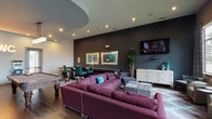 This game room is a well-appointed recreational space designed to accommodate a variety of social and leisure activities. The layout is thoughtfully divided into distinct zones, each serving a specific purpose while maintaining a cohesive aesthetic throughout.

The billiards area serves as a central focal point, anchored by a full-size pool table with dark wood legs and gray felt, positioned beneath a geometric chandelier with cube-shaped fixtures. A wall-mounted cue rack and a large flat-screen TV are positioned on the adjacent blue-gray accent wall, making this corner ideal for game viewing and play alike.

The lounge area features generous sectional seating in deep plum upholstery, accented with teal throw pillows, arranged over a patterned area rug in lavender and aqua tones. A white lattice-door credenza serves as a media console beneath a wall-mounted TV, flanked by framed artwork and a tall potted plant. Additional seating includes gray accent chairs and ottoman cubes for flexible arrangements.

A high-top table with teal chairs provides a dedicated spot for card games or casual dining. Large floor-to-ceiling windows along one wall flood the space with natural light, offering outdoor views. Dark hardwood flooring and layered accent walls in charcoal and slate blue lend the space a polished, contemporary ambiance.