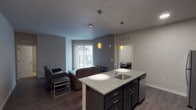 This single-floor unit offers a well-designed, functional layout suited for multiple occupants. The open-concept kitchen and living area serves as the heart of the space, where the kitchen stands out with dark espresso cabinetry, granite countertops, stainless steel appliances including a refrigerator, range, and microwave, and a central island with a built-in sink. Stylish pendant lighting with amber glass shades adds a warm, contemporary touch overhead. Light wood-style flooring runs throughout the kitchen and living area, while the living space opens to a sliding glass door with vertical blinds, allowing natural light to fill the room and providing access to an outdoor view. Multiple bedrooms feature neutral-toned walls, carpeted floors, individual windows with blinds, and sufficient space for a bed and desk. The unit includes two full bathrooms, each finished with a white vanity, large mirror, and a walk-in shower stall equipped with a built-in bench seat and chrome fixtures — one of which features accessibility-friendly grab bars. The overall aesthetic is clean and modern, with a neutral color palette and practical finishes throughout.