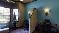 This single-floor space presents a refined and thoughtfully designed interior that combines rich color and classic architectural details to create a warm, professional ambiance. The walls are dressed in a striking combination of deep teal and warm neutral tones, framed by dark wood crown molding that adds a polished, sophisticated touch throughout. Hardwood flooring runs underfoot, contributing a sense of warmth and continuity to the space. Wall sconces with cream-shaded fixtures provide soft, ambient lighting that complements the overall atmosphere, while a ceiling-mounted light fixture adds additional illumination from above. A dedicated workspace is positioned near a window dressed with a rich red valance, allowing natural light to filter into the room and offering views of lush greenery outside. A dark wood half-wall partition adds architectural interest and helps define zones within the open layout. Decorative elements, including a potted plant near the window and tasteful accents on a dark wood side table, enhance the character of the space. The interior strikes a balance between functionality and elegance, making it well-suited for both focused work and welcoming visitors.