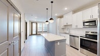 This well-appointed 2-bedroom, 2-bathroom residence combines modern design with functional living across a single-story open floor plan. The heart of the home is a stylish open-concept kitchen and living area featuring light wood-style flooring, recessed lighting, and a large central island with a crisp white quartz countertop, illuminated by two globe pendant lights. The kitchen is outfitted with white shaker-style cabinetry, stainless steel appliances including a dishwasher and range, and a stainless steel sink. Natural light pours in through large windows and a glass-paneled entry door, creating a bright and airy atmosphere that extends into the living area. Both bedrooms are generously sized, offering neutral-toned walls, light wood-style flooring, and windows that bring in natural light. The primary bedroom is notably spacious and is served by a private bathroom featuring a walk-in glass shower with white subway tile and direct access to a dedicated walk-in closet with built-in shelving and hanging rods. The second bathroom includes a full soaking tub and shower combo with white subway tile, a white vanity with quartz countertop, and access to an additional closet with built-in organization. Clean lines, neutral tones, and contemporary finishes give the entire residence a cohesive, modern aesthetic.