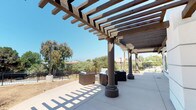 Located at 912 3rd Avenue in Chula Vista, California, this single-story outdoor space is defined by a substantial wood-beam pergola structure that extends along a wide, smooth concrete patio. The pergola is supported by sturdy, decorative columns with classic base detailing, casting a distinctive latticed shadow pattern across the patio surface on sunny days — a hallmark of Southern California's abundant natural light.

The patio is generously proportioned, offering ample room for multiple seating arrangements. Outdoor furniture, including modular sofas and accent chairs, is positioned beneath the pergola's shade, creating a comfortable setting for gatherings or relaxation. The space is bordered by decorative iron fencing, which frames views of mature trees and natural landscaping beyond the perimeter.

Access to the adjacent interior is provided through full-height glass-paneled double doors set into a clean, white stucco exterior wall, connecting indoor and outdoor spaces seamlessly. On one side of the patio, a children's playground is visible, featuring green play structures and soft ground cover, adding a family-friendly dimension to the overall environment. The surrounding neighborhood's residential buildings are visible in the background, grounding the space within its community context.