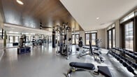 This single-level fitness center is designed to support a comprehensive workout experience, combining thoughtful architectural details with a wide array of professional-grade equipment. The space is divided into distinct functional zones, each tailored to different aspects of physical training.

The main workout area features a striking wood-paneled ceiling that adds warmth and visual interest, contrasting with the light-toned walls and polished epoxy flooring below. Ceiling fans are strategically positioned to maintain airflow throughout. Recessed lighting supplements the abundant natural light that streams in through large, floor-to-ceiling windows and paneled glass doors, offering views of an outdoor pool area and surrounding grounds.

Strength training is well-represented with a multi-station cable machine, free-weight benches, a full dumbbell rack, a heavy punching bag, and various resistance machines. A dedicated cardio zone houses treadmills, an elliptical, and a stair climber, all positioned near mirrored walls that enhance the sense of space. Wall-mounted televisions are installed throughout for entertainment during workouts. A plyo box and additional functional training accessories round out the facility's offerings.